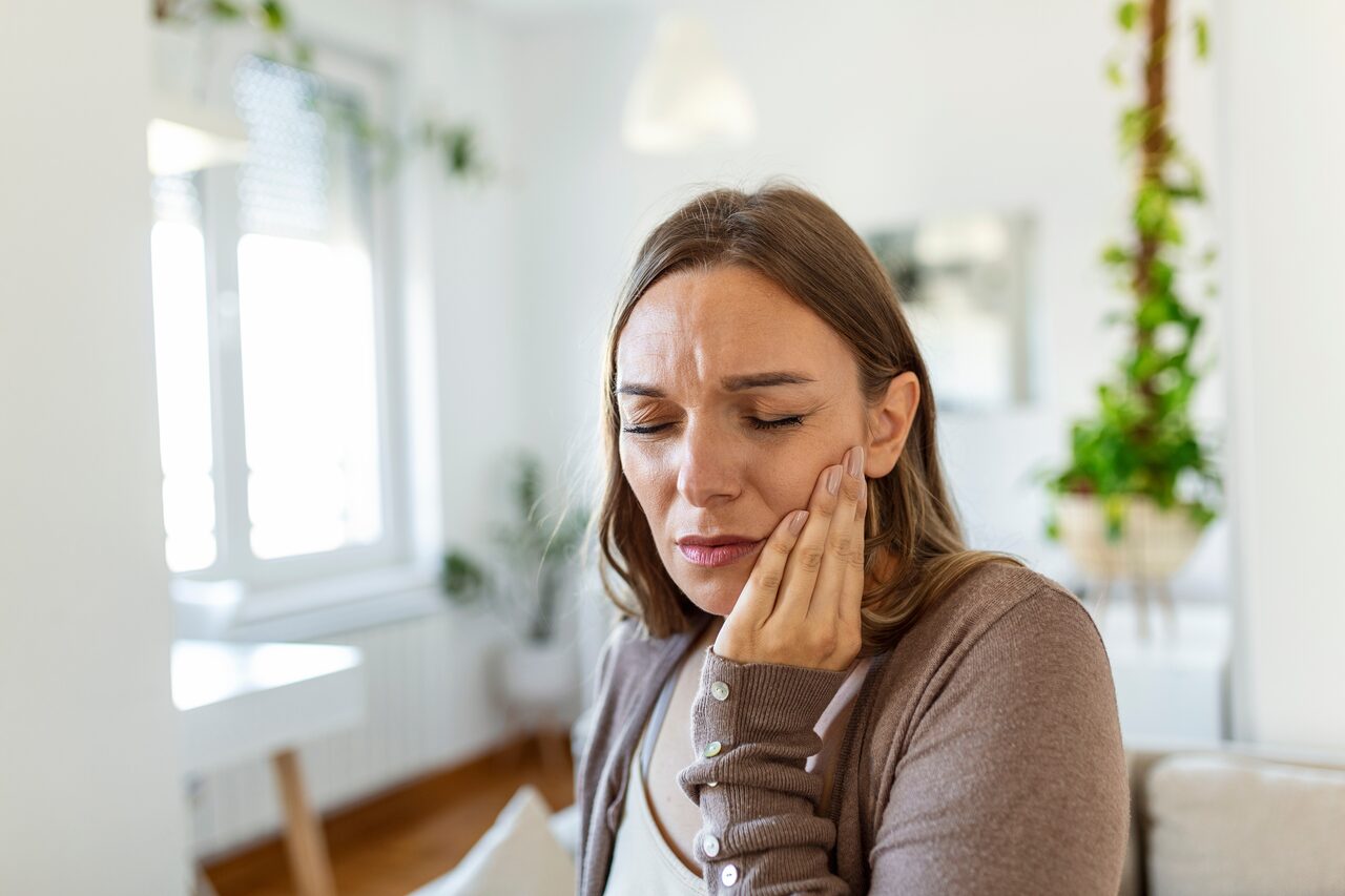 A woman at home wincing and holding her cheek from jaw pain